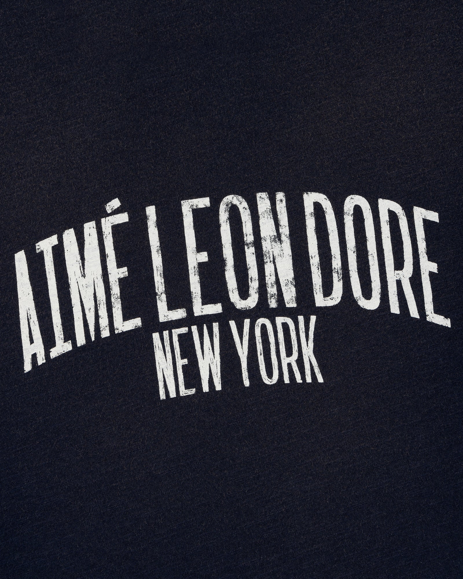 White, distressed "AIMÉ LEON DORE NEW YORK" text on the Washed Ringer Tee's dark fabric background.