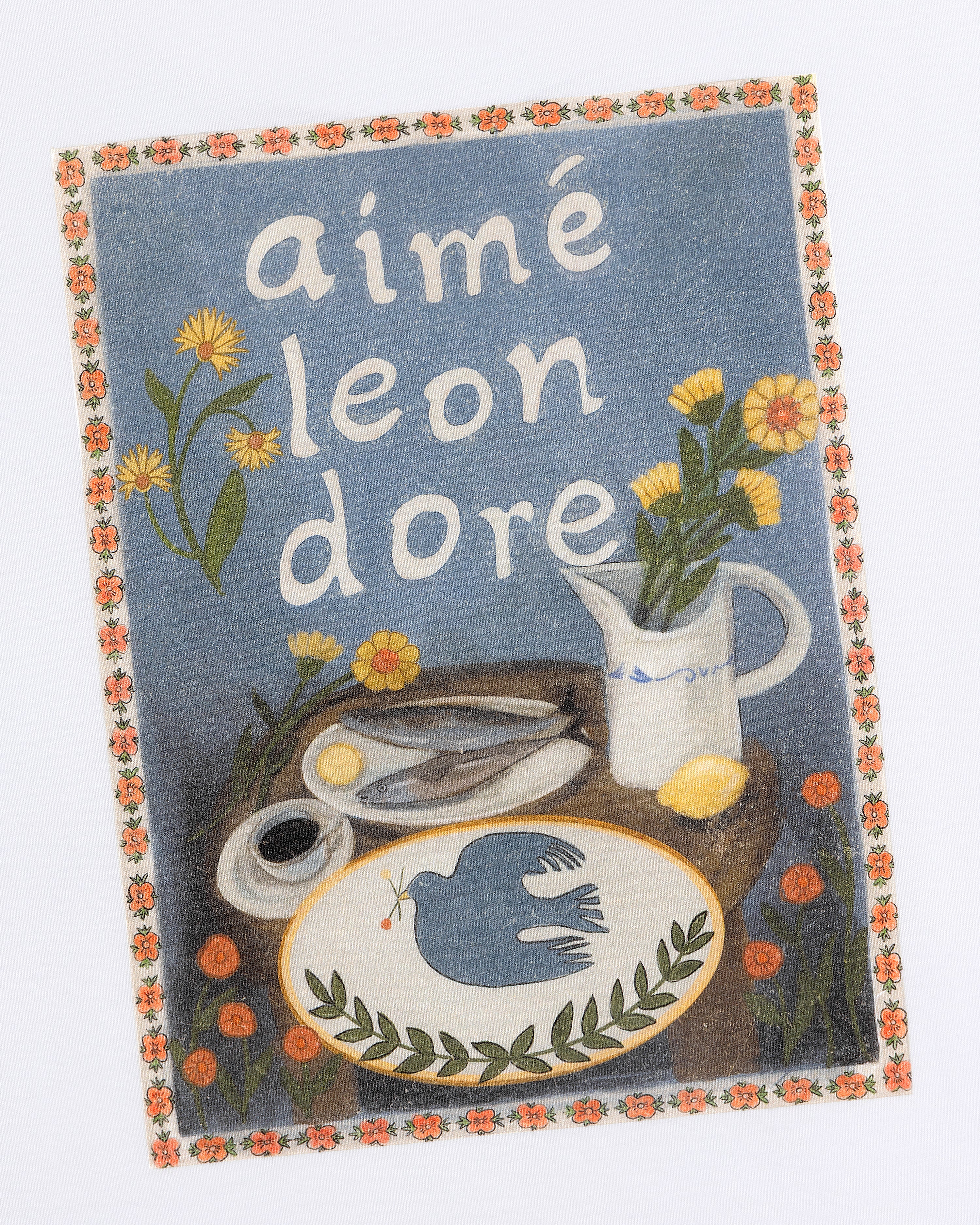 Breakfast Tee features a floral border with flowers, a bird, pitcher, scissors, lemon, and Aimé Leon Dore on a table.
