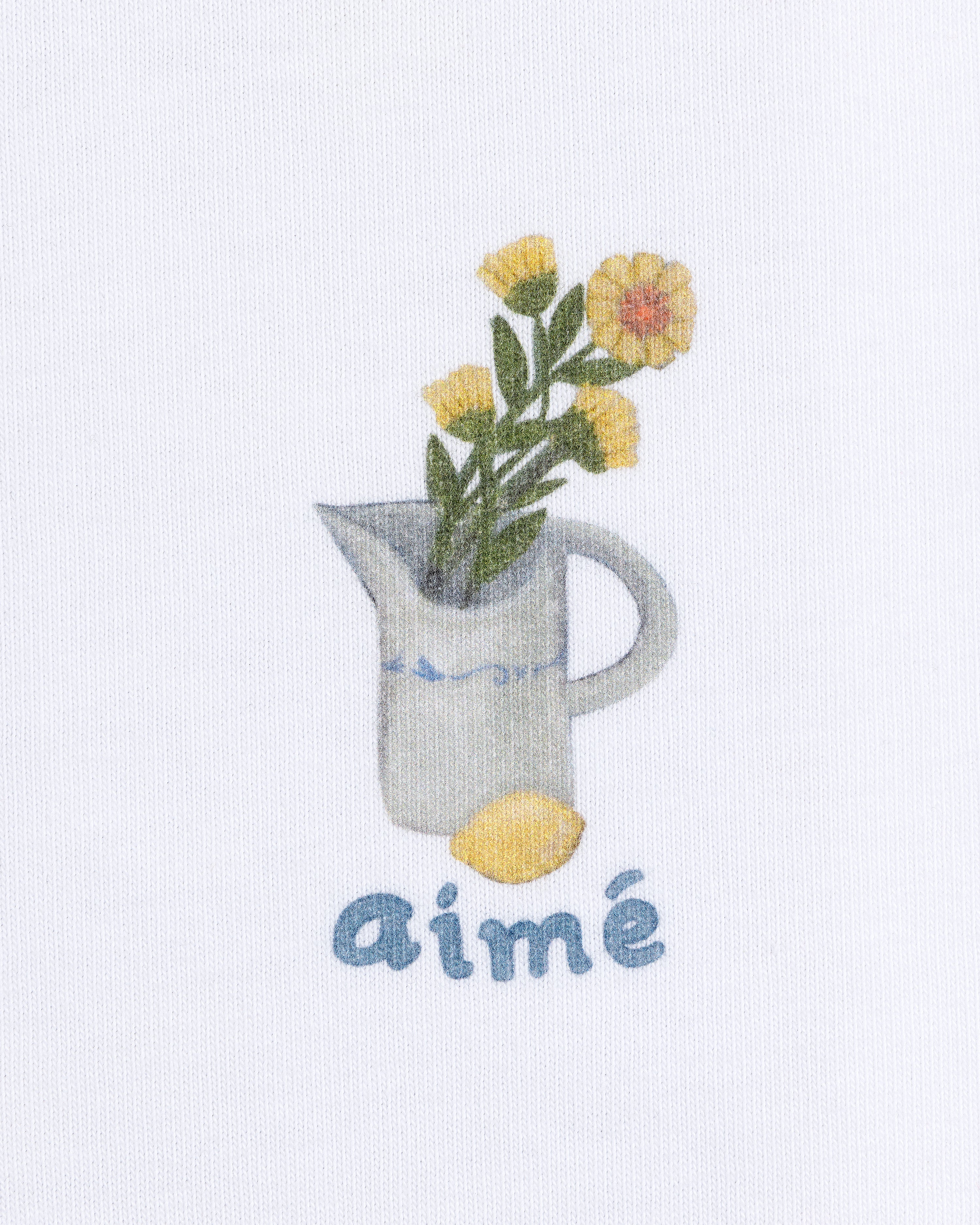 The Breakfast Tee features a white pitcher with yellow flowers, a lemon, and "aimé" in blue text below.