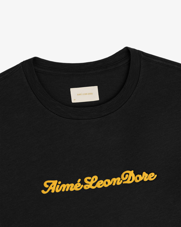 Black Puff Print Slub Logo Tee featuring Aimé Leon Dore in yellow cursive across the chest.