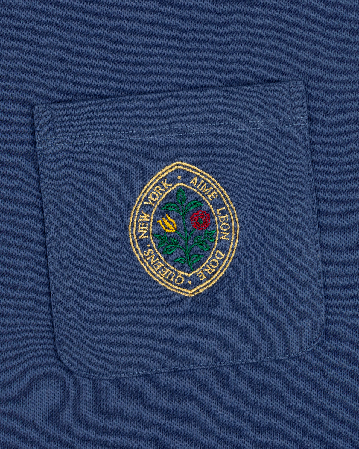 Botanical Crest Pocket Tee: blue shirt pocket with a circular floral patch and "New York - Ames Leon Dore - Queens.