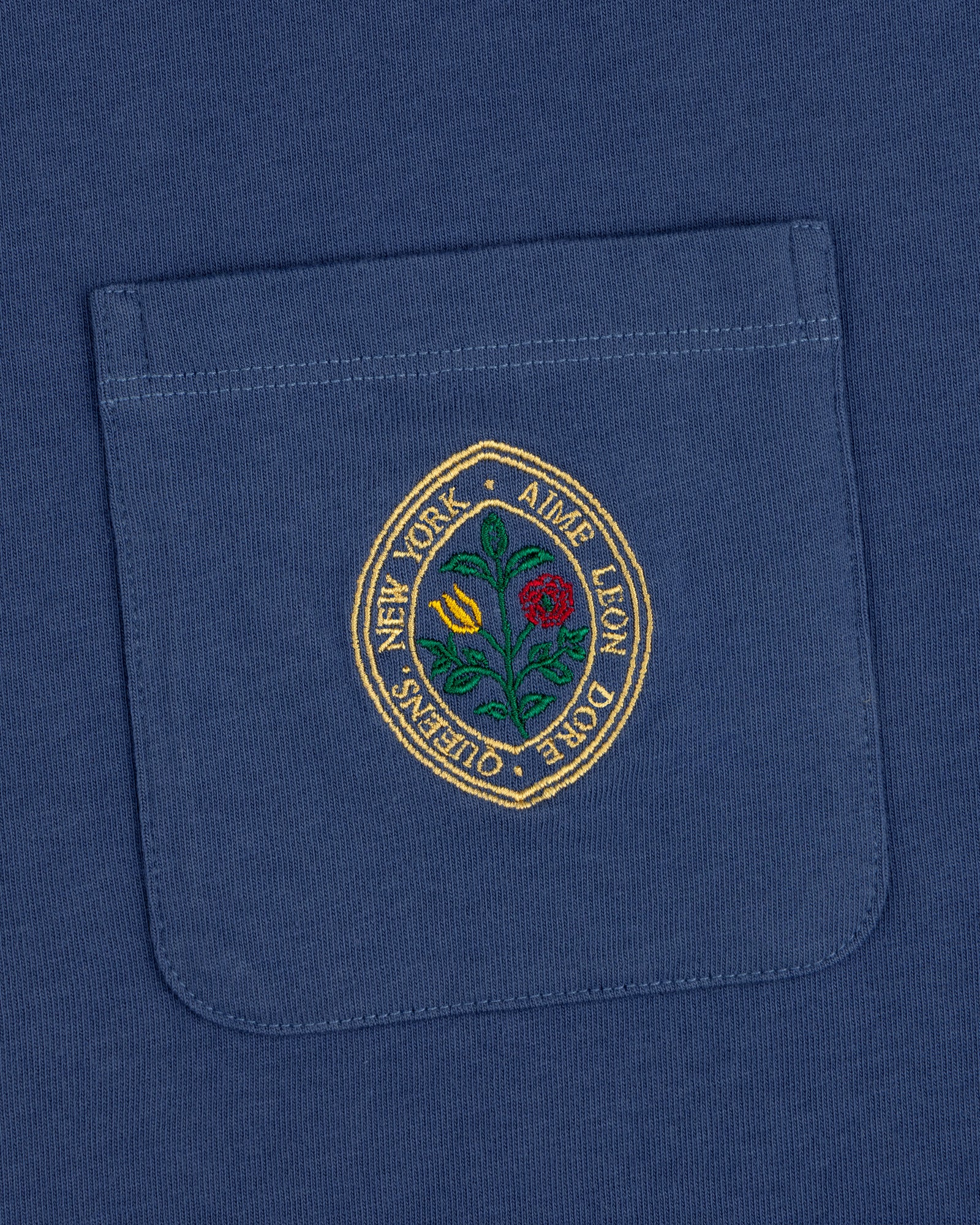 Botanical Crest Pocket Tee: blue shirt pocket with a circular floral patch and "New York - Ames Leon Dore - Queens.