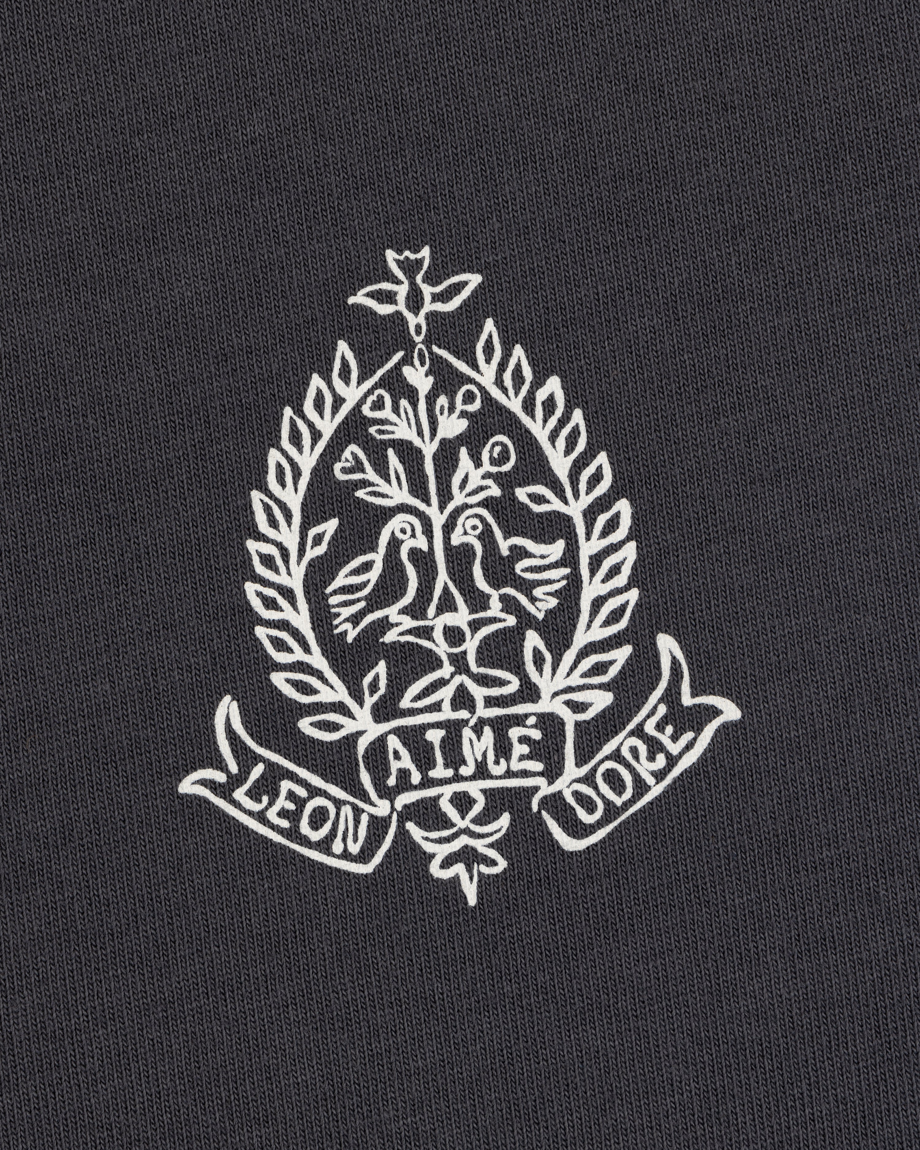 White embroidery of two birds, leafy branches, and "LEON AIME DOPE" on the Saint George Graphic Tee.