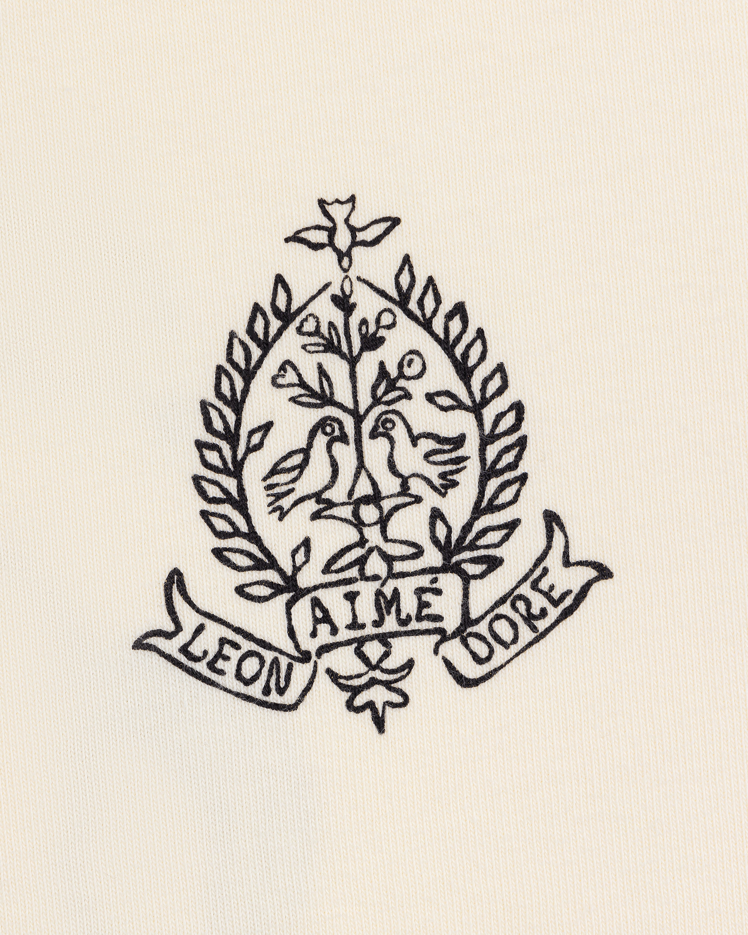 Black line art of two birds on branches in a laurel wreath, with LEON and DORE banners, on the Saint George Graphic Tee.