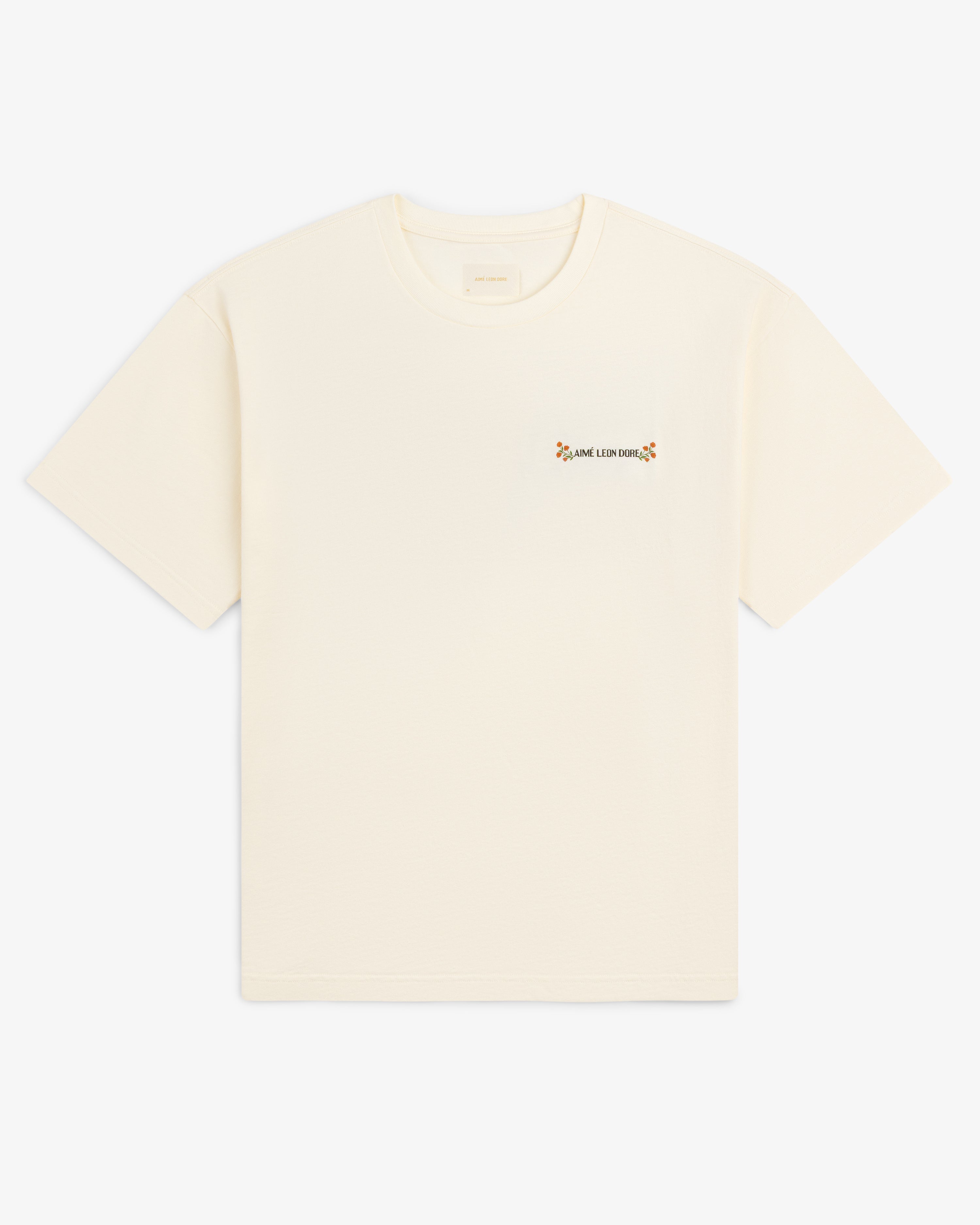 Aimé Postcard Tee: off-white short-sleeve shirt with small floral graphic and text on the left chest.