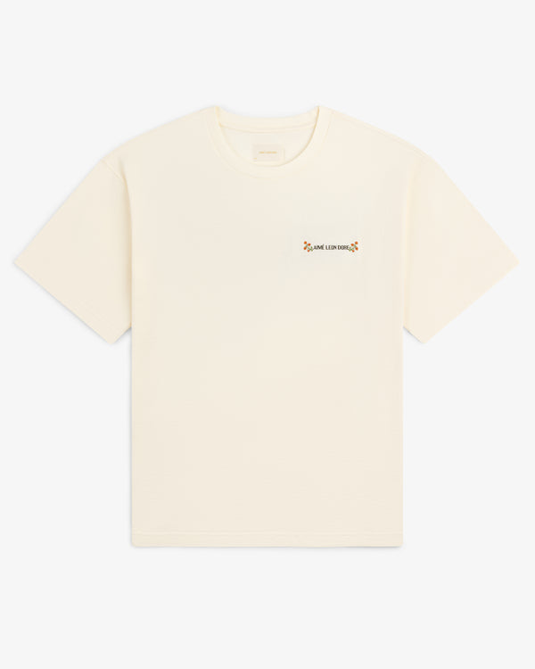 Aimé Postcard Tee: off-white short-sleeve shirt with small floral graphic and text on the left chest.