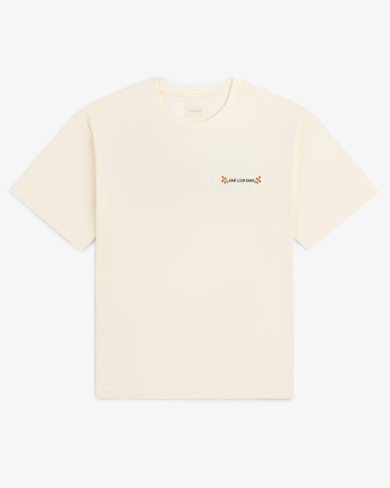 Aimé Postcard Tee: off-white short-sleeve shirt with small floral graphic and text on the left chest.