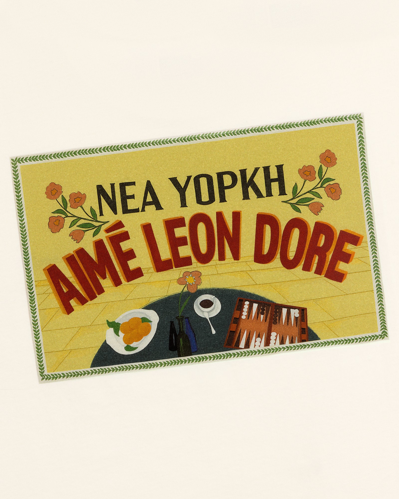 Rectangular patch on Aimé Postcard Tee with NEA YOPKH AIMÉ LEON DORE, flowers, food, cup, and books.