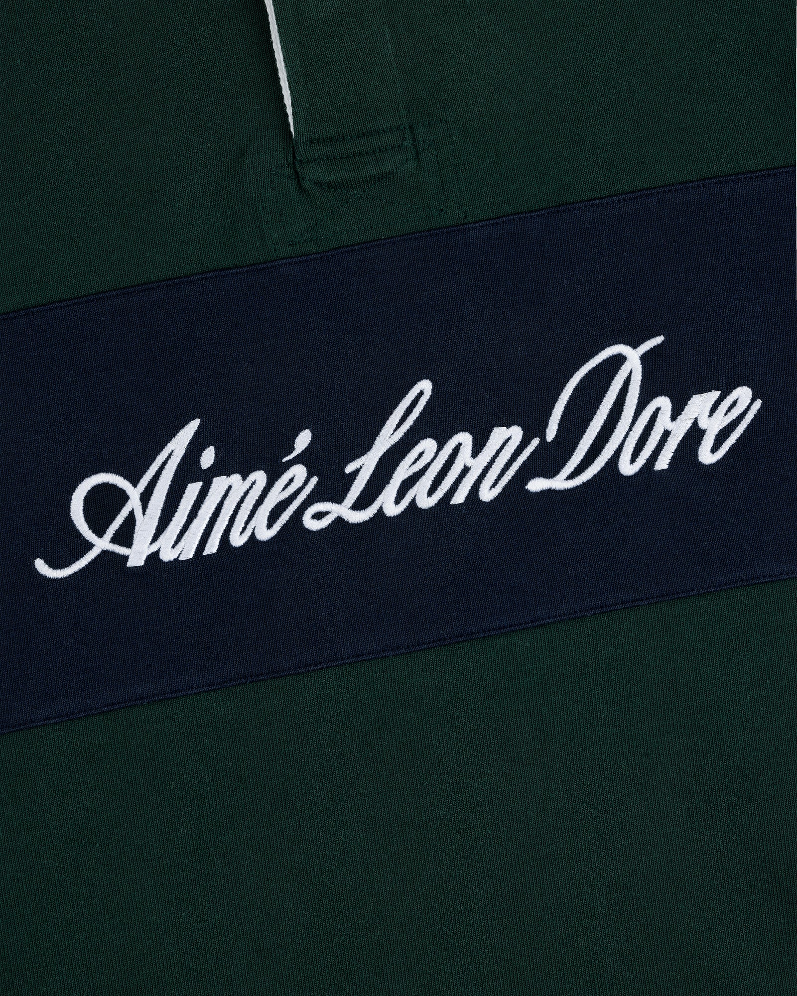 Close-up of Script Paneled Rugby: dark green fabric with a navy stripe and Aimé Leon Dore in white script.