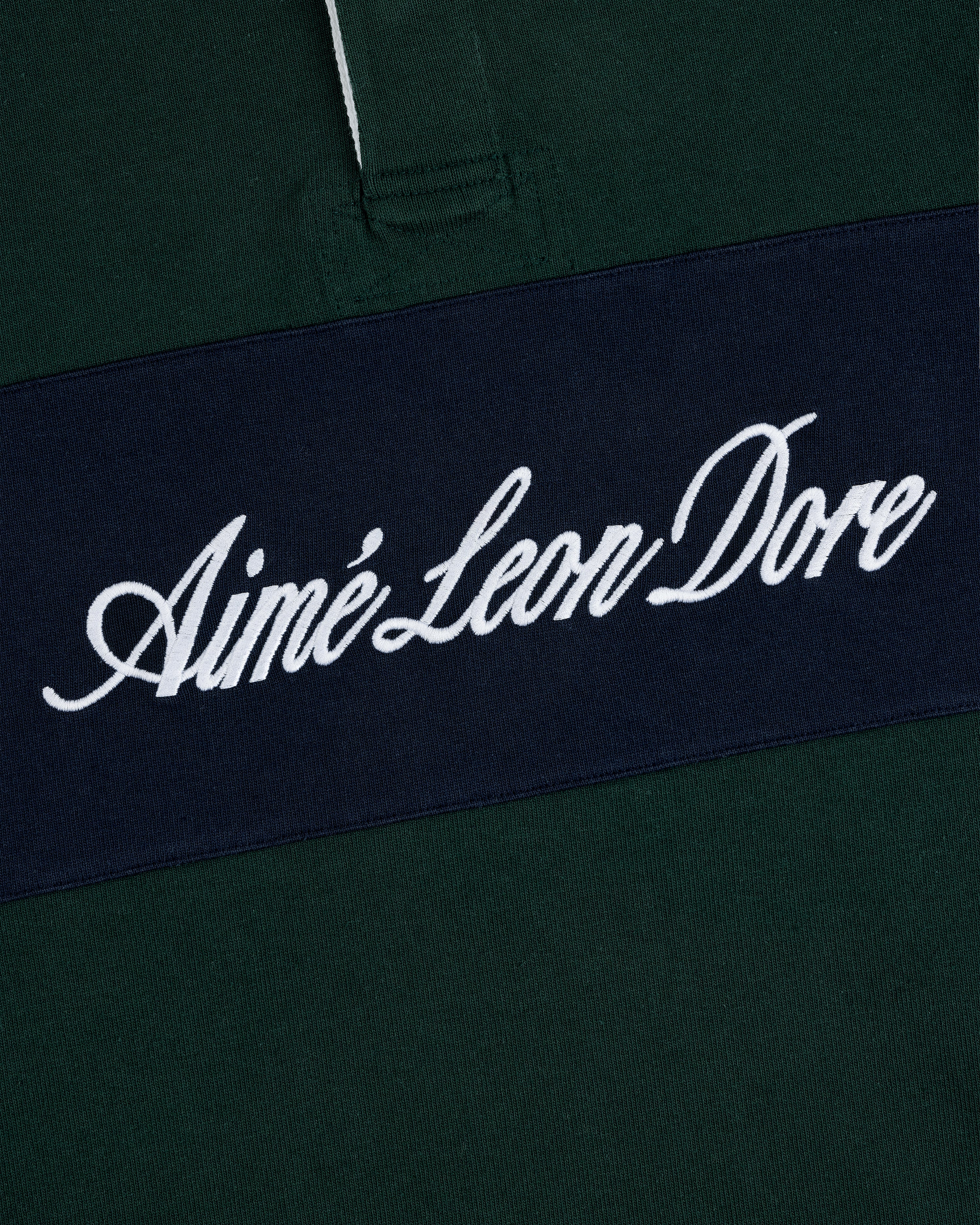 Close-up of Script Paneled Rugby: dark green fabric with a navy stripe and Aimé Leon Dore in white script.