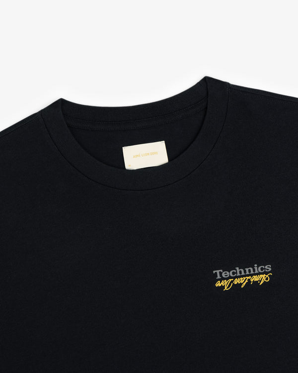 Black ALD / Technics Logo Tee with small yellow script on left chest; beige collar tag visible.