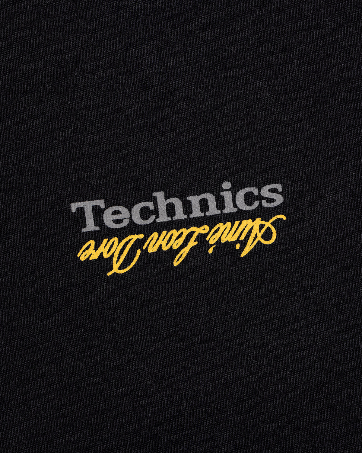 The ALD / Technics Logo Tee features gray "Technics" and yellow aline icon dore printed upside down on black fabric.