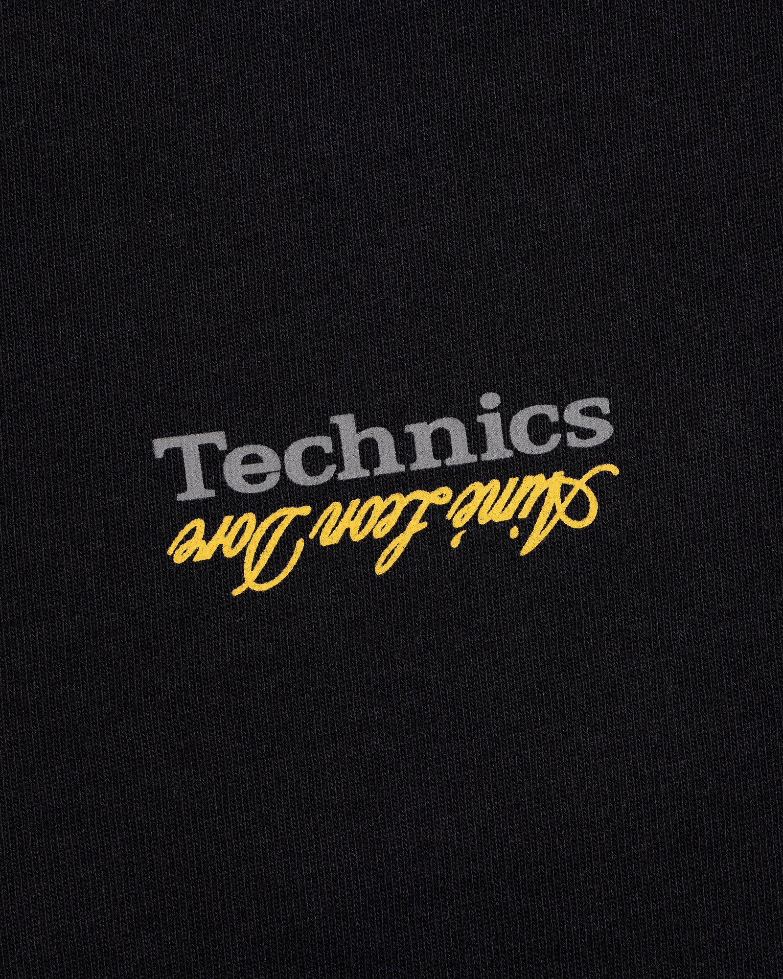 The ALD / Technics Logo Tee features gray "Technics" and yellow aline icon dore printed upside down on black fabric.