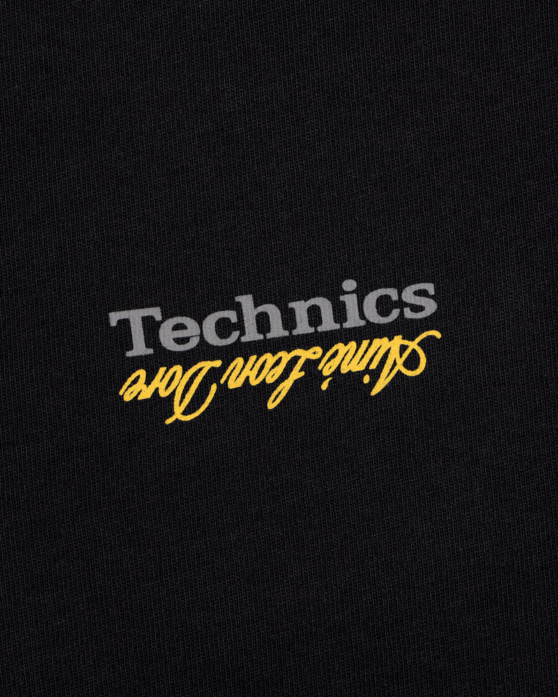 The ALD / Technics Logo Tee features gray "Technics" and yellow aline icon dore printed upside down on black fabric.