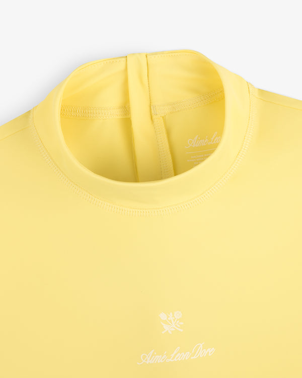 Close-up of a yellow Short Sleeve Crest Rash Guard with a white logo and floral graphic on the chest.