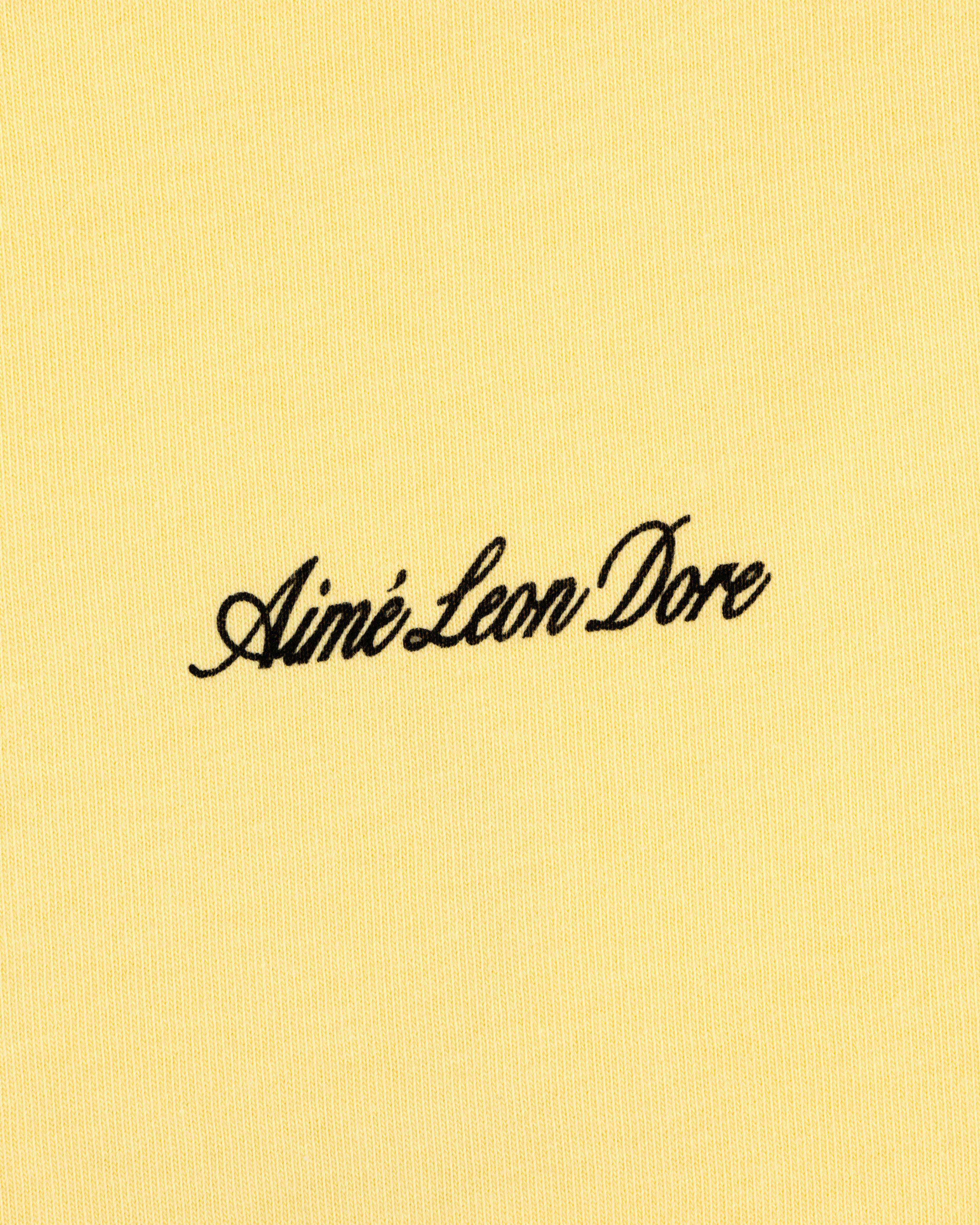 Yellow Super Cub Tee with Aimé Leon Dore in black cursive text at the center.