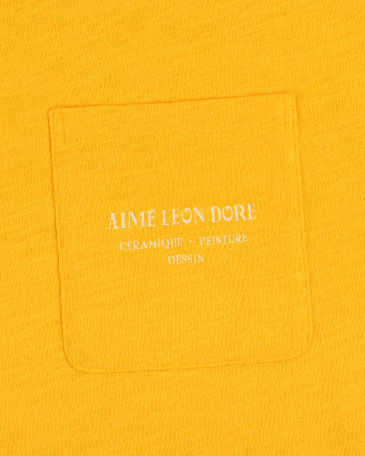 Artiste Pocket Tee in yellow features a pocket with white text: Aime Leon Dore Céramique - Peinture Dessin.