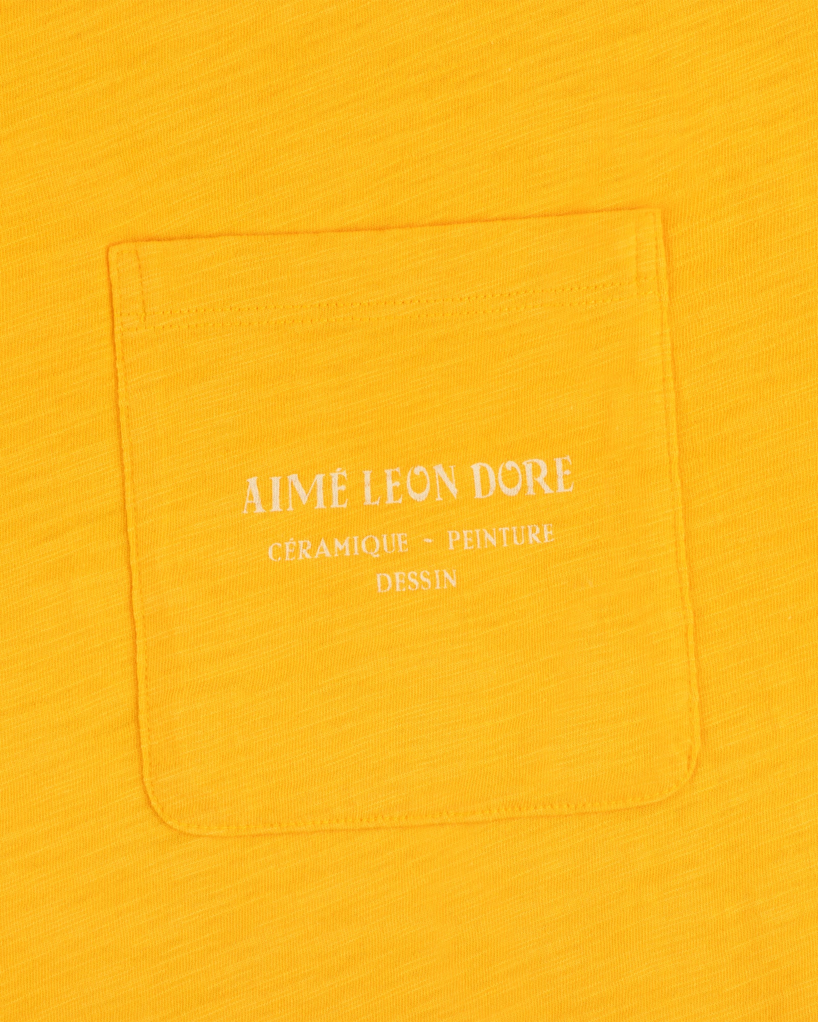 Artiste Pocket Tee in yellow features a pocket with white text: Aime Leon Dore Céramique - Peinture Dessin.