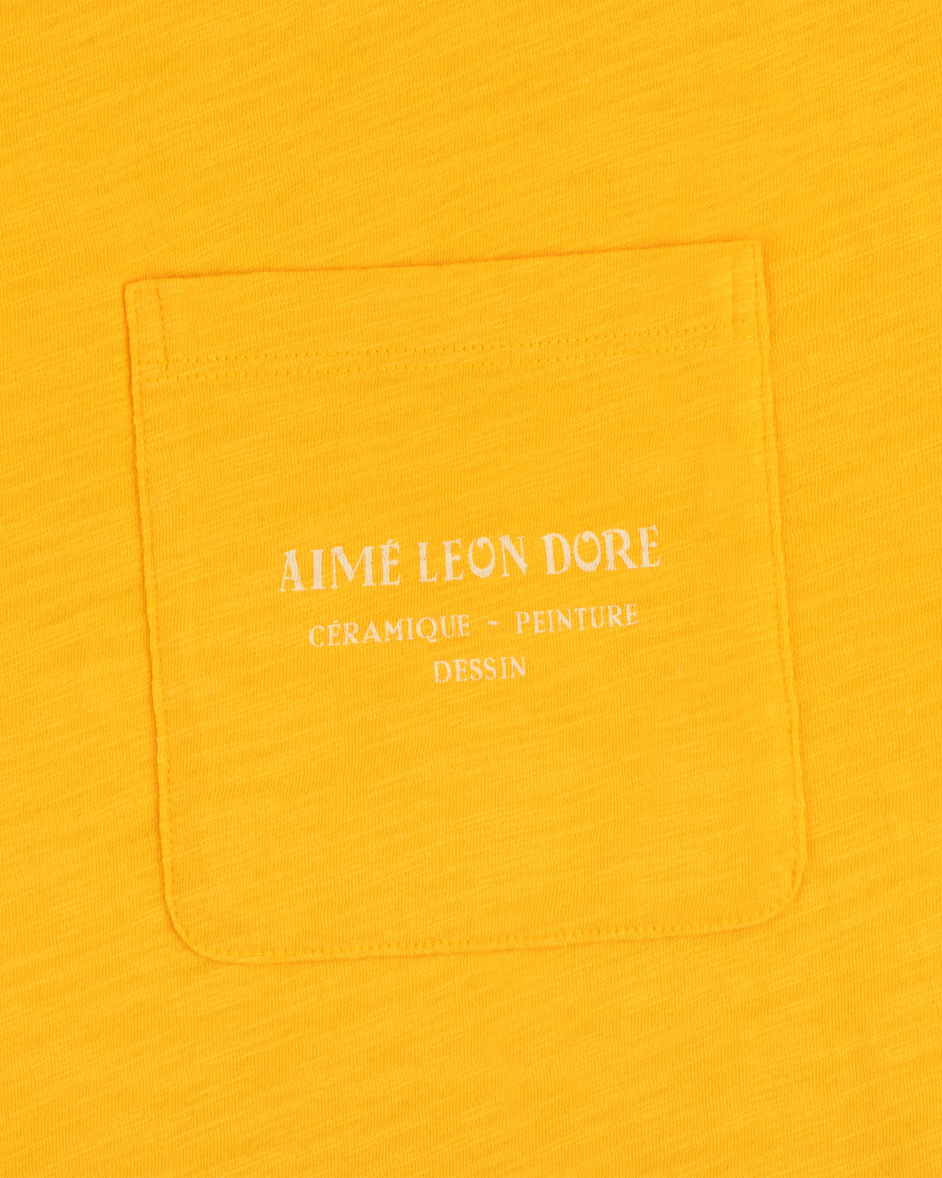 Artiste Pocket Tee in yellow features a pocket with white text: Aime Leon Dore Céramique - Peinture Dessin.
