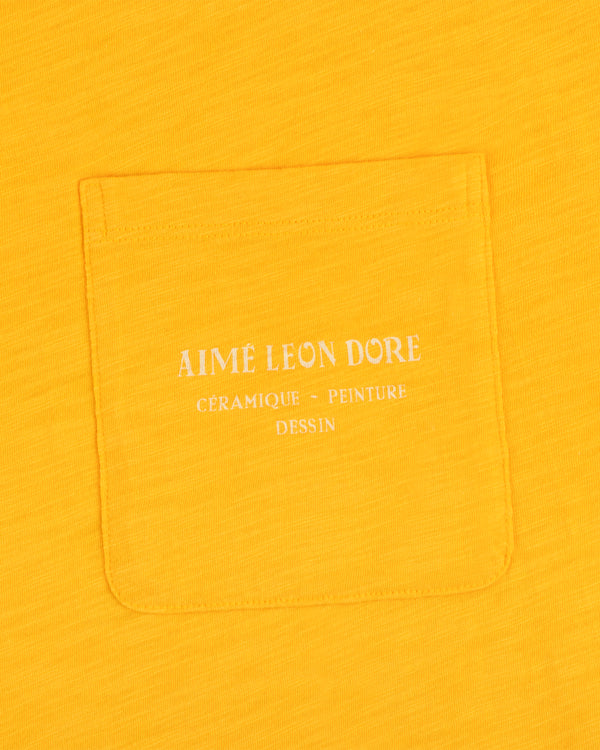 Artiste Pocket Tee in yellow features a pocket with white text: Aime Leon Dore Céramique - Peinture Dessin.