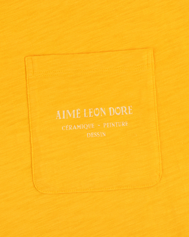 Artiste Pocket Tee in yellow features a pocket with white text: Aime Leon Dore Céramique - Peinture Dessin.