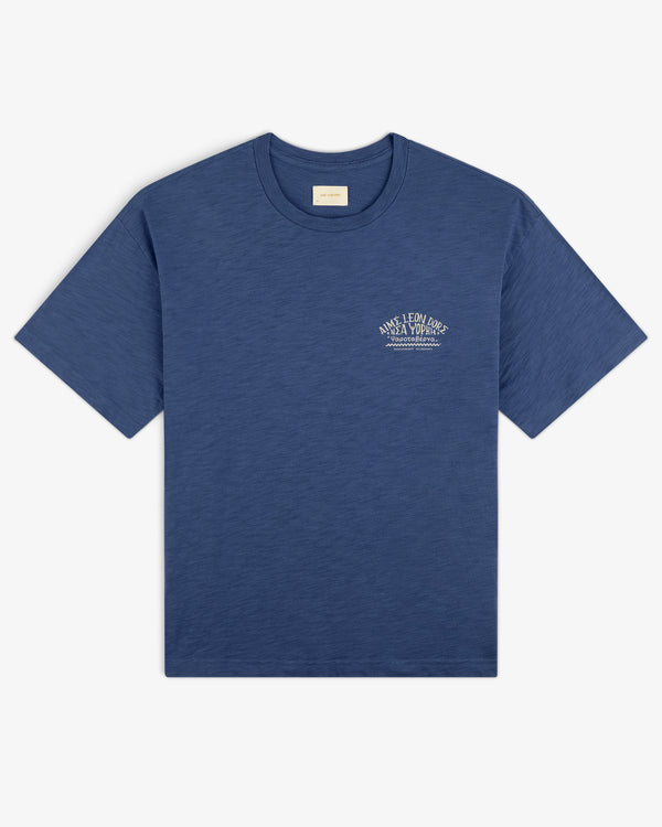 Taverna Tee in blue, short sleeves, with small white text logo on left chest, laid flat on white background.