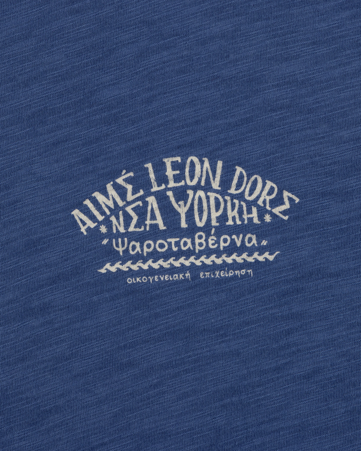Greek and English text printed on blue Taverna Tee, featuring "AIMÉ LEON DORE NEW YORK.