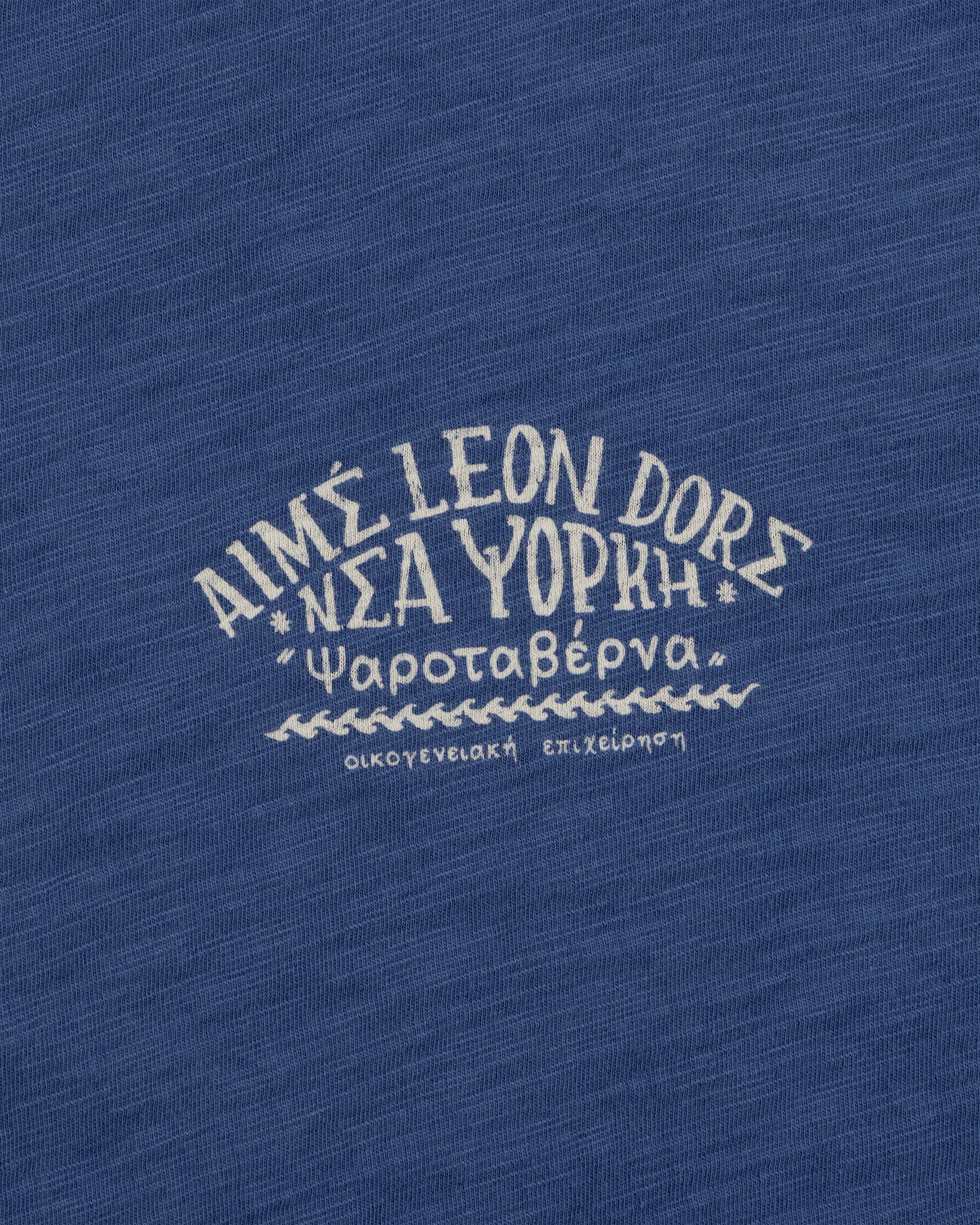 Greek and English text printed on blue Taverna Tee, featuring "AIMÉ LEON DORE NEW YORK.