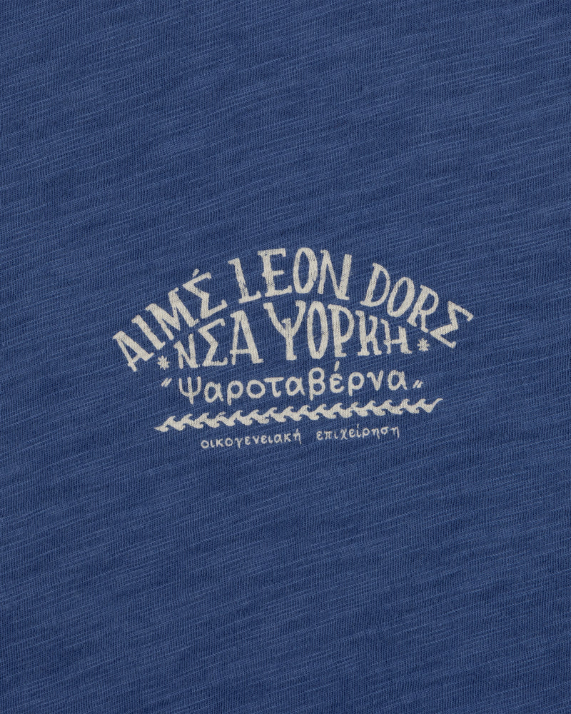 Greek and English text printed on blue Taverna Tee, featuring "AIMÉ LEON DORE NEW YORK.