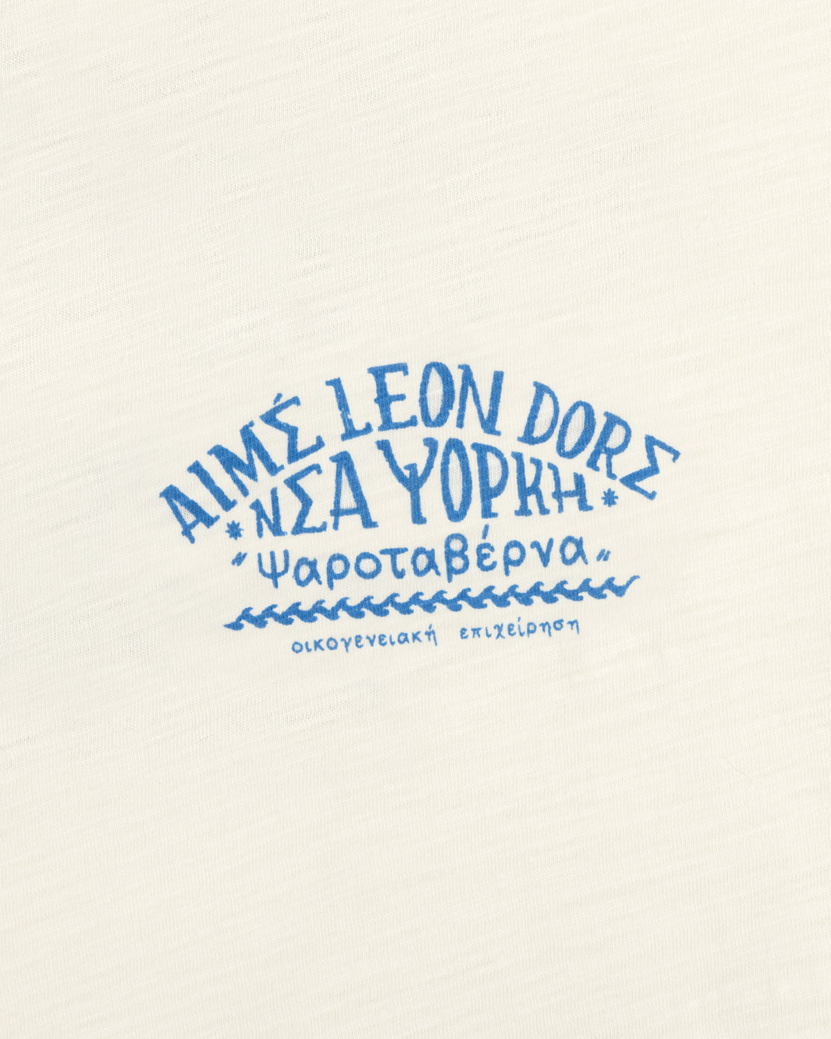 White Taverna Tee with blue Greek & English text: AIMÉ LEON DORE ΝΕΑ ΥΟΡΚΗ Ψαροταβέρνα οικογενειακή επιχείρηση.