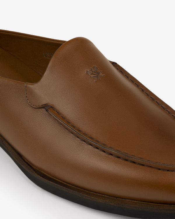 Close-up of the Venetian Mule, a brown leather loafer with visible stitching and an embossed logo on the upper.