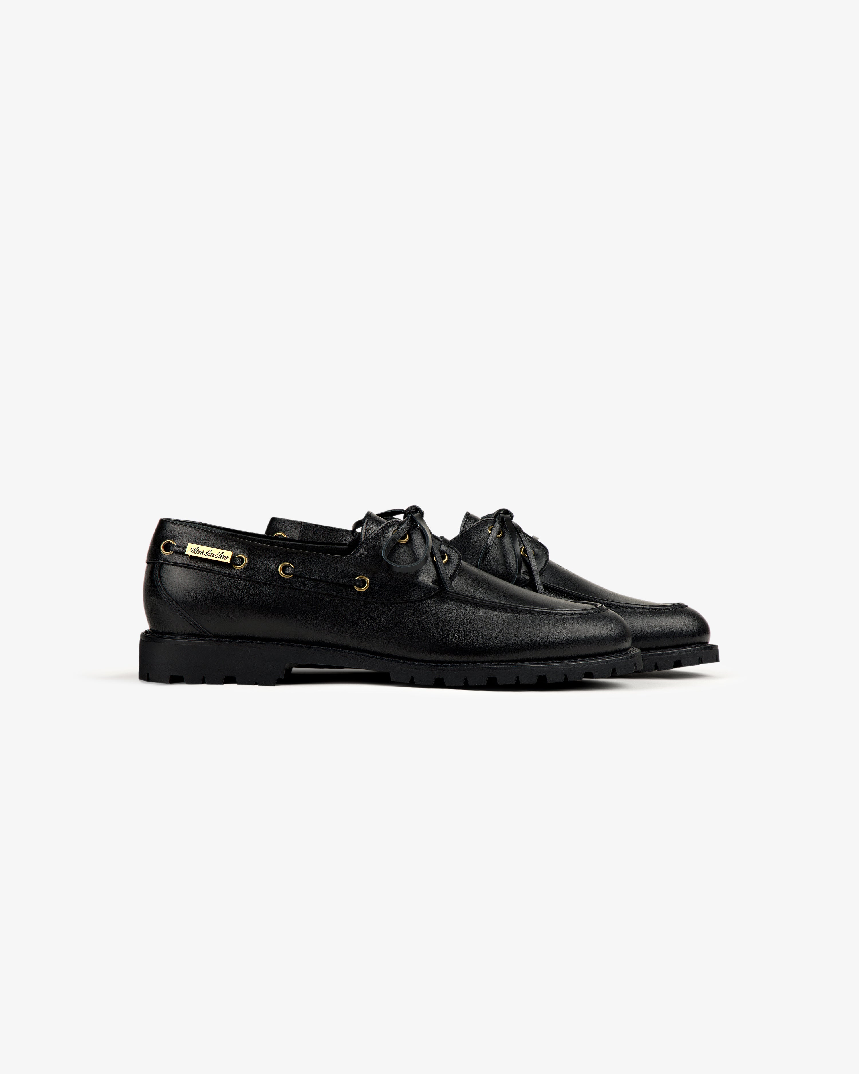 The Pápia Boat Shoe: black leather lace-up with gold eyelets and chunky rubber soles, shown on a white background.