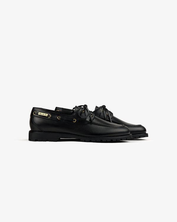 The Pápia Boat Shoe: black leather lace-up with gold eyelets and chunky rubber soles, shown on a white background.