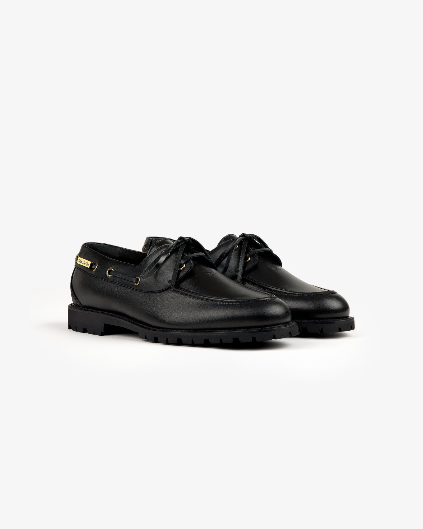 The Pápia Boat Shoe: black leather lace-ups with gold eyelets and thick rubber soles on a white background.