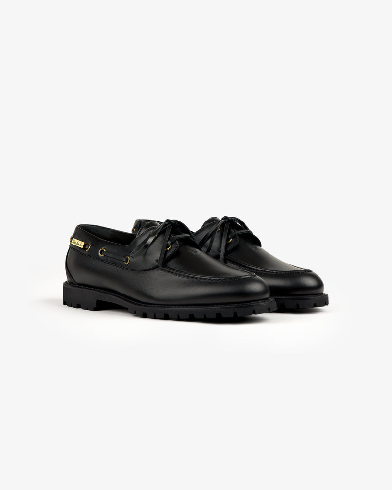 The Pápia Boat Shoe: black leather lace-ups with gold eyelets and thick rubber soles on a white background.