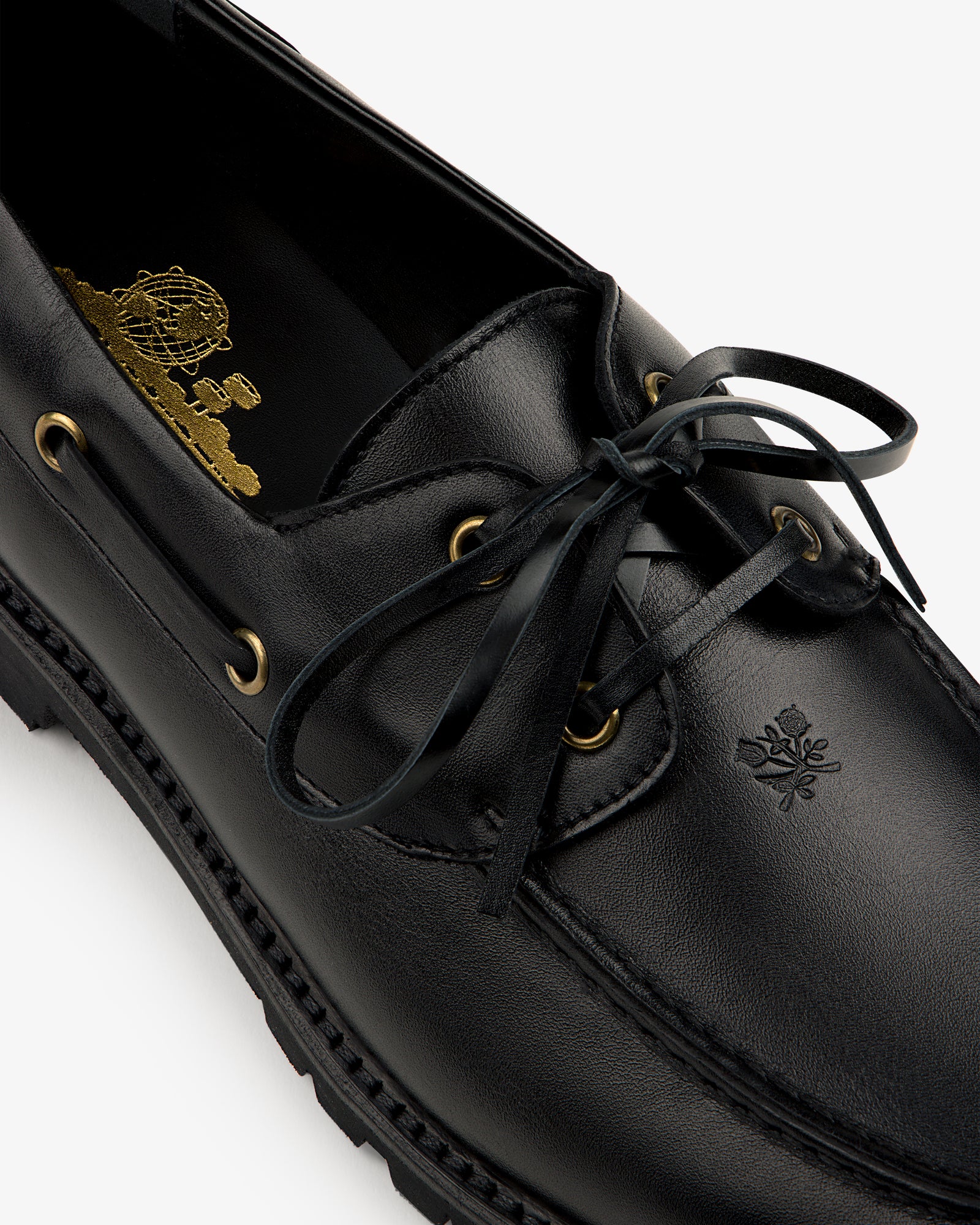 Close-up of the Pápia Boat Shoe: black leather loafer with gold eyelets, black laces, and an embossed toe crest.