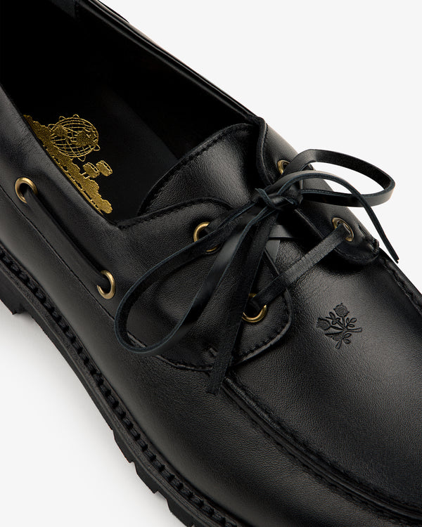 Close-up of the Pápia Boat Shoe: black leather loafer with gold eyelets, black laces, and an embossed toe crest.