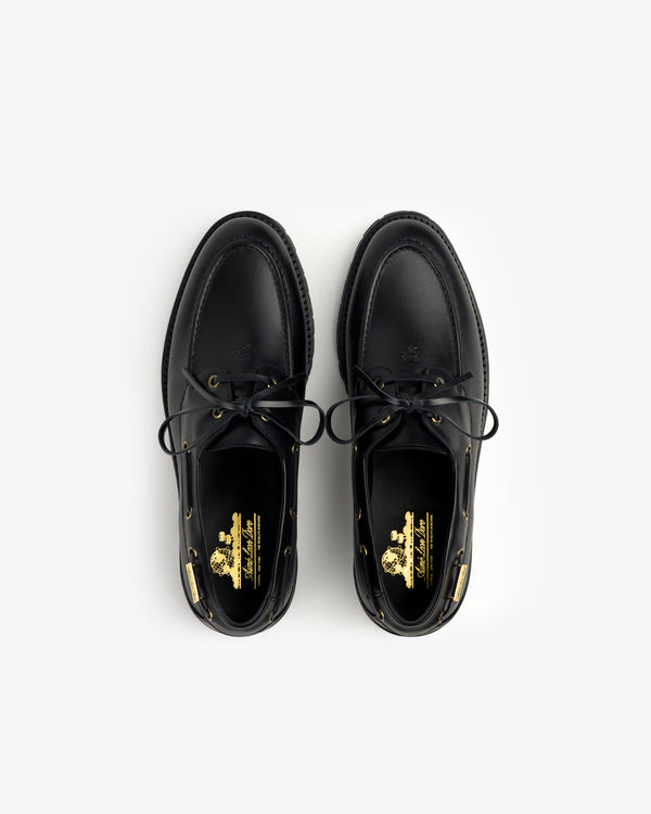 The Pápia Boat Shoe is a black leather lace-up shoe with gold logo details on the insoles, seen from above.