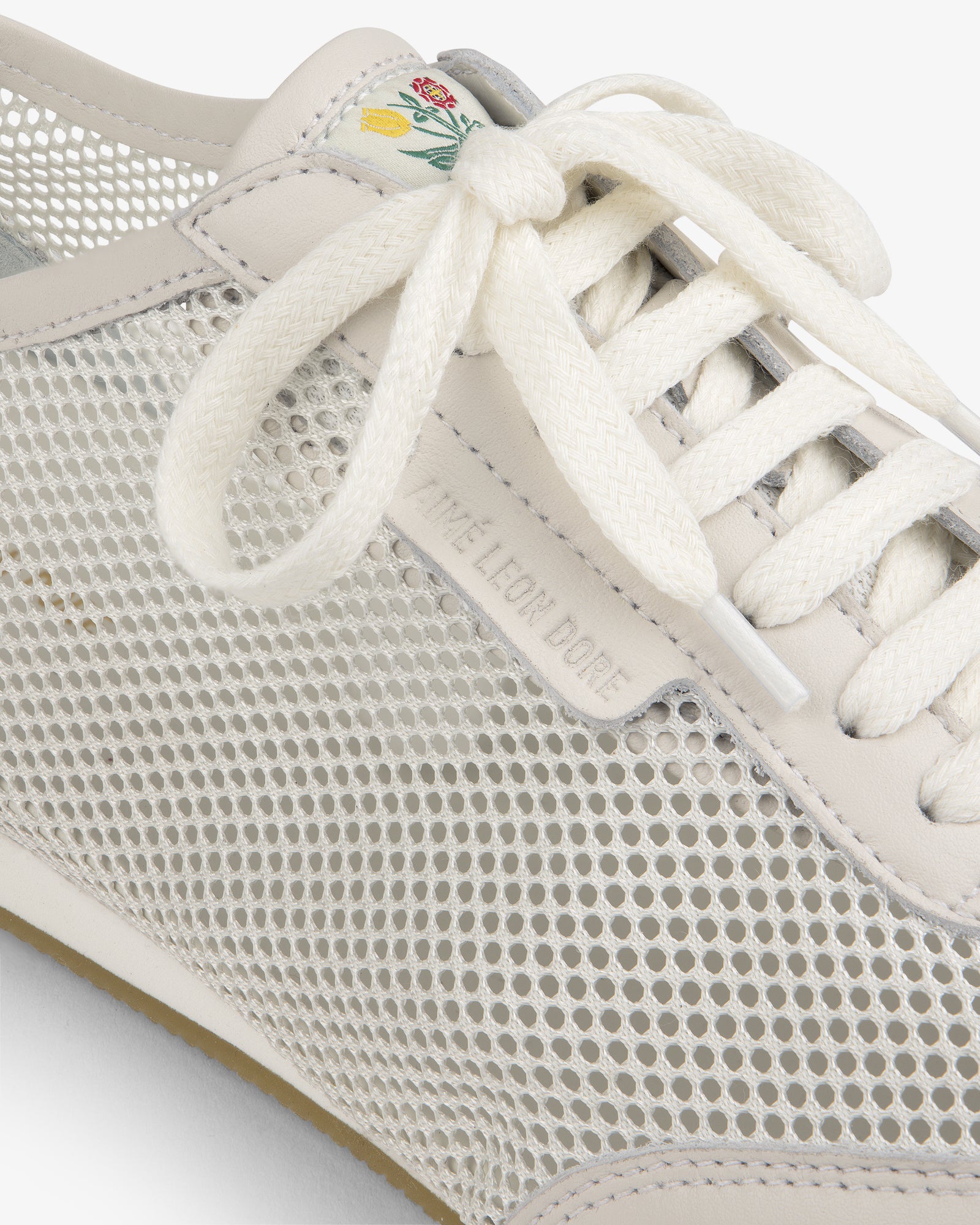 Close-up of the Low Profile Mesh Trainer, a white mesh sneaker with cream laces and a small flower on the tongue.