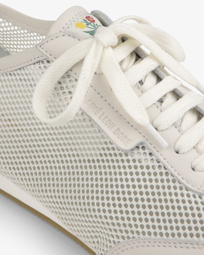 Close-up of the Low Profile Mesh Trainer, a white mesh sneaker with cream laces and a small flower on the tongue.