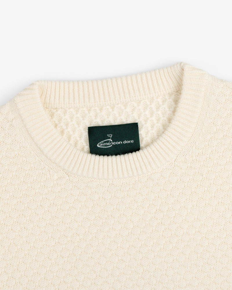 Close-up of the ALD Golf Clubhouse Sweater, a cream knit with a ribbed crew neck and green Aimé Leon Dore label.
