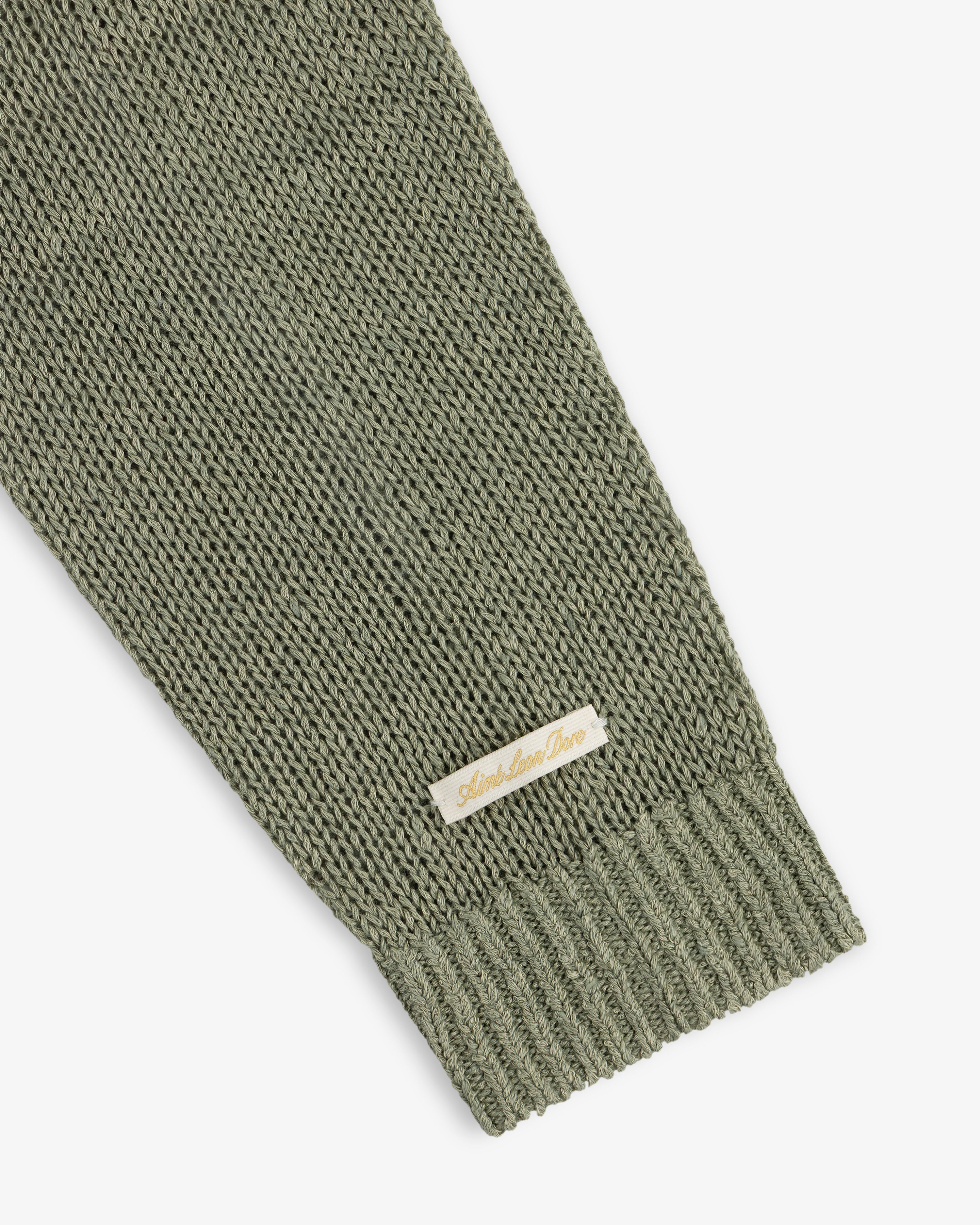 A close-up of the Linen Knit Crewneck Sweater sleeve in olive green, featuring a small beige label near the cuff.
