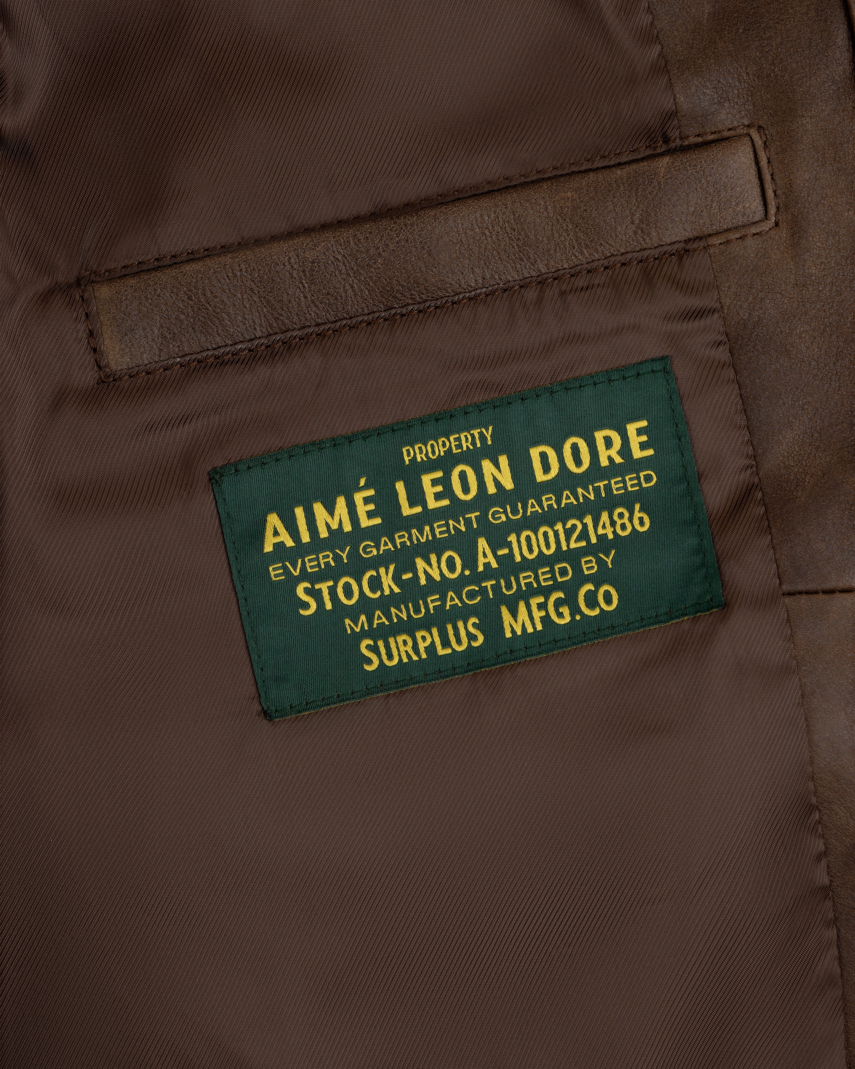 Close-up of a green Aimé Leon Dore label sewn inside the World's Borough Leather Flight Jacket on brown fabric.