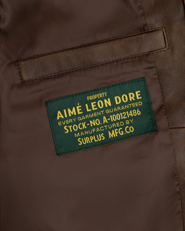 Close-up of a green Aimé Leon Dore label sewn inside the World's Borough Leather Flight Jacket on brown fabric.