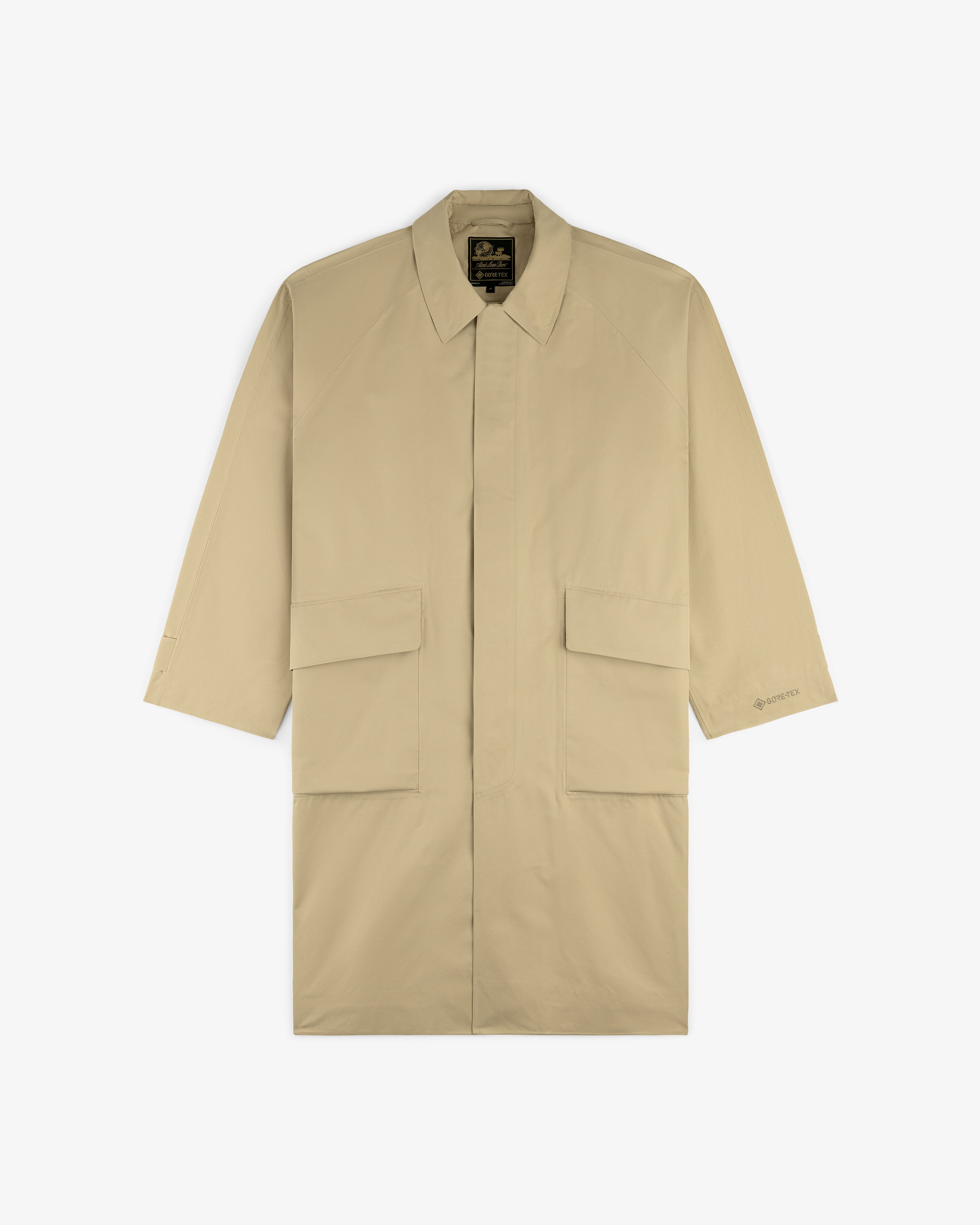 The GORE-TEX Balmacaan Coat is a beige, long-sleeve trench with large front flap pockets and a collar, laid flat.