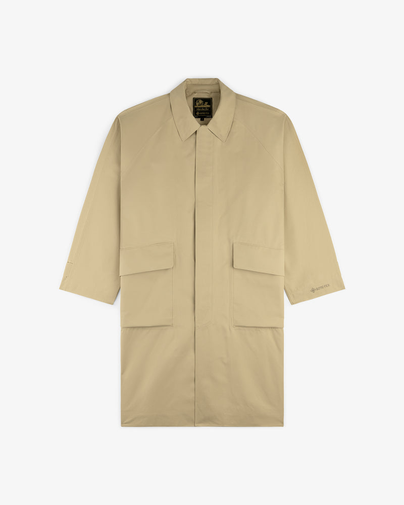 The GORE-TEX Balmacaan Coat is a beige, long-sleeve trench with large front flap pockets and a collar, laid flat.