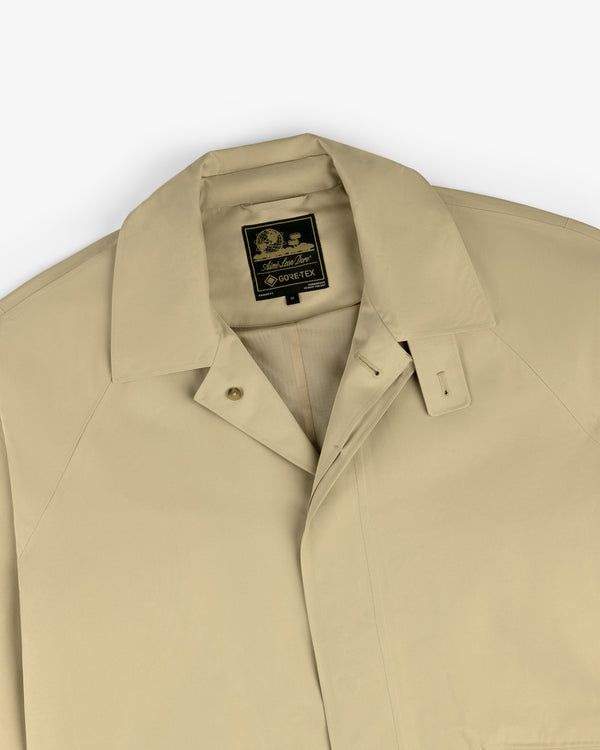 The GORE-TEX Balmacaan Coat is beige, collared, and features a black and gold interior label, shown top front.