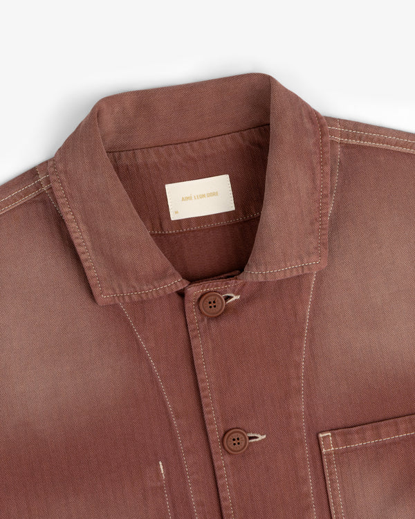 Rust-colored Sun Faded Chore Jacket with white stitching, two brown buttons, and ARNE LOMBRIKE label inside collar.