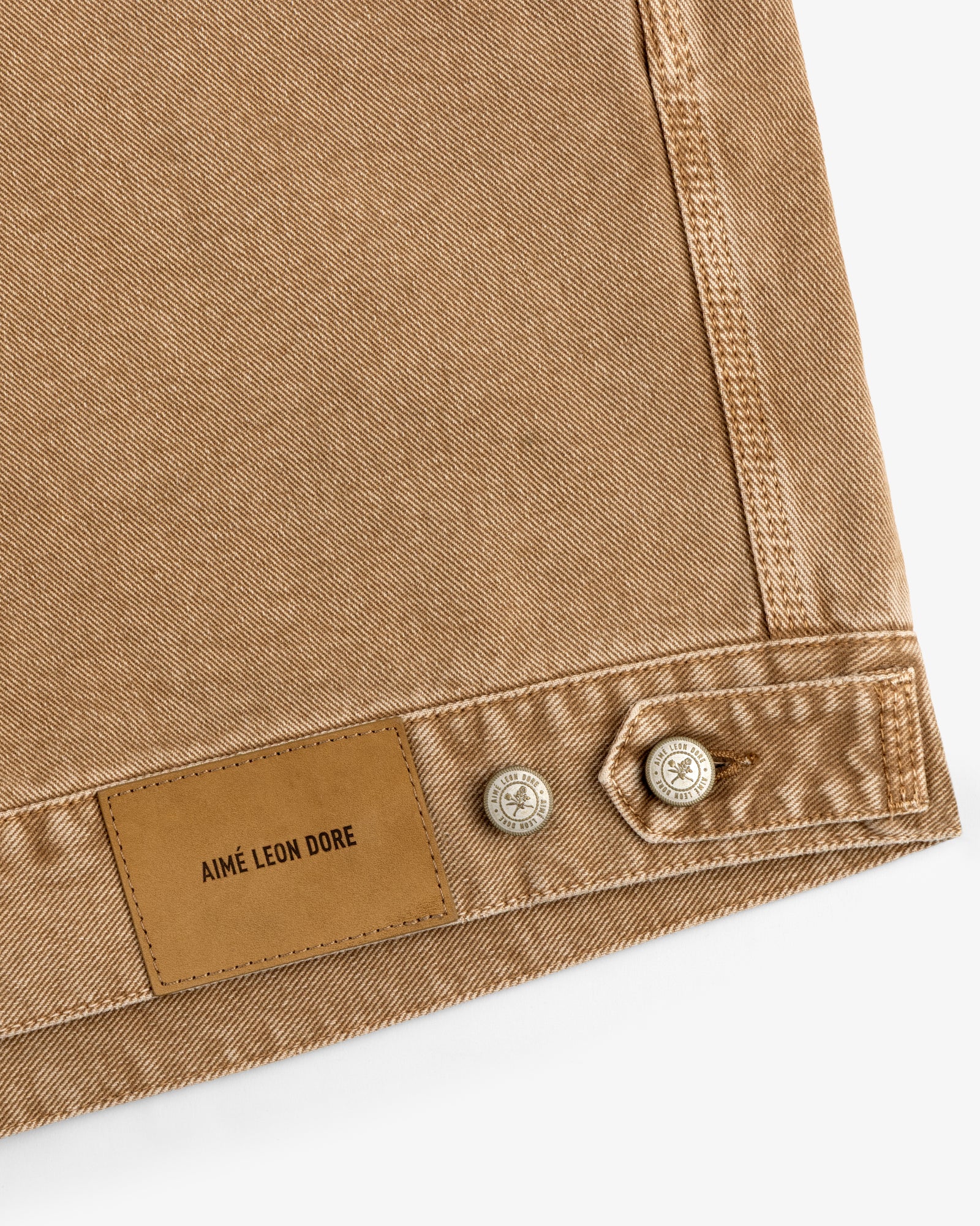 Close-up of tan Denim Trucker Jacket showing a leather Aimé Leon Dore label and two metal hem buttons.