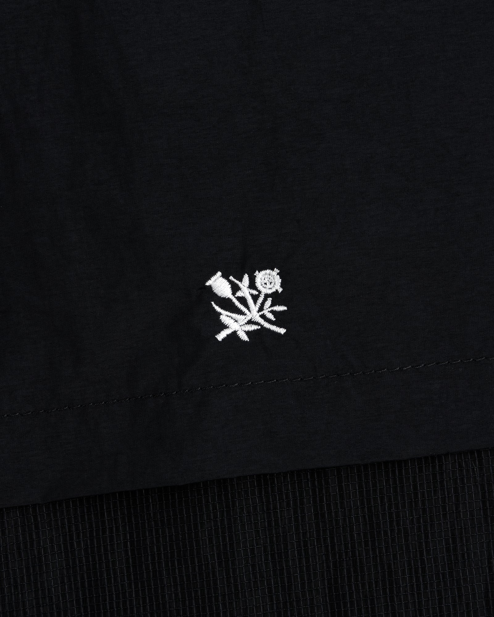 Small white embroidered rose with leaves on black Nylon Track Jacket, near the hem.